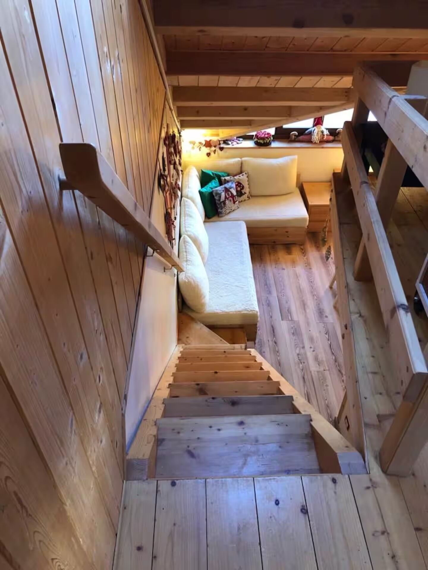 Una scala in legno conduce a una zona con divano, cuscini e un piccolo tavolino, il tutto circondato da pareti e una ringhiera in legno.