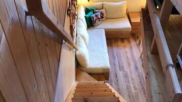 Una scala in legno conduce a una zona con divano, cuscini e un piccolo tavolino, il tutto circondato da pareti e una ringhiera in legno.