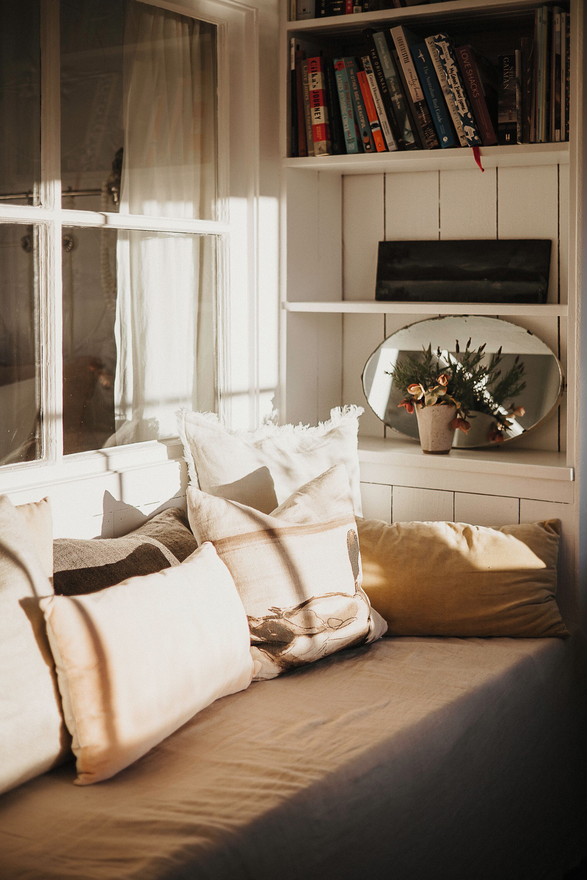 A cozy reading nook with a window, a bookshelf, and a mirror reflecting a plant.