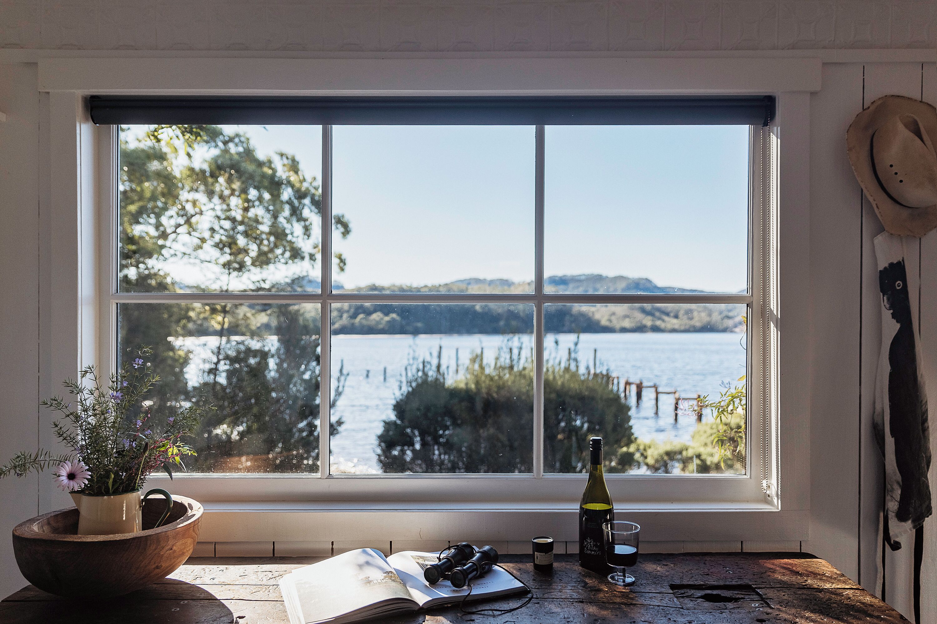 A room with a large window overlooking a lake, a wooden table with a wine bottle, glasses, and a book, and a potted plant on the left.