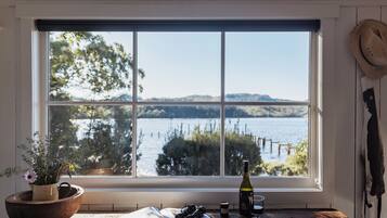 A room with a large window overlooking a lake, a wooden table with a wine bottle, glasses, and a book, and a potted plant on the left.