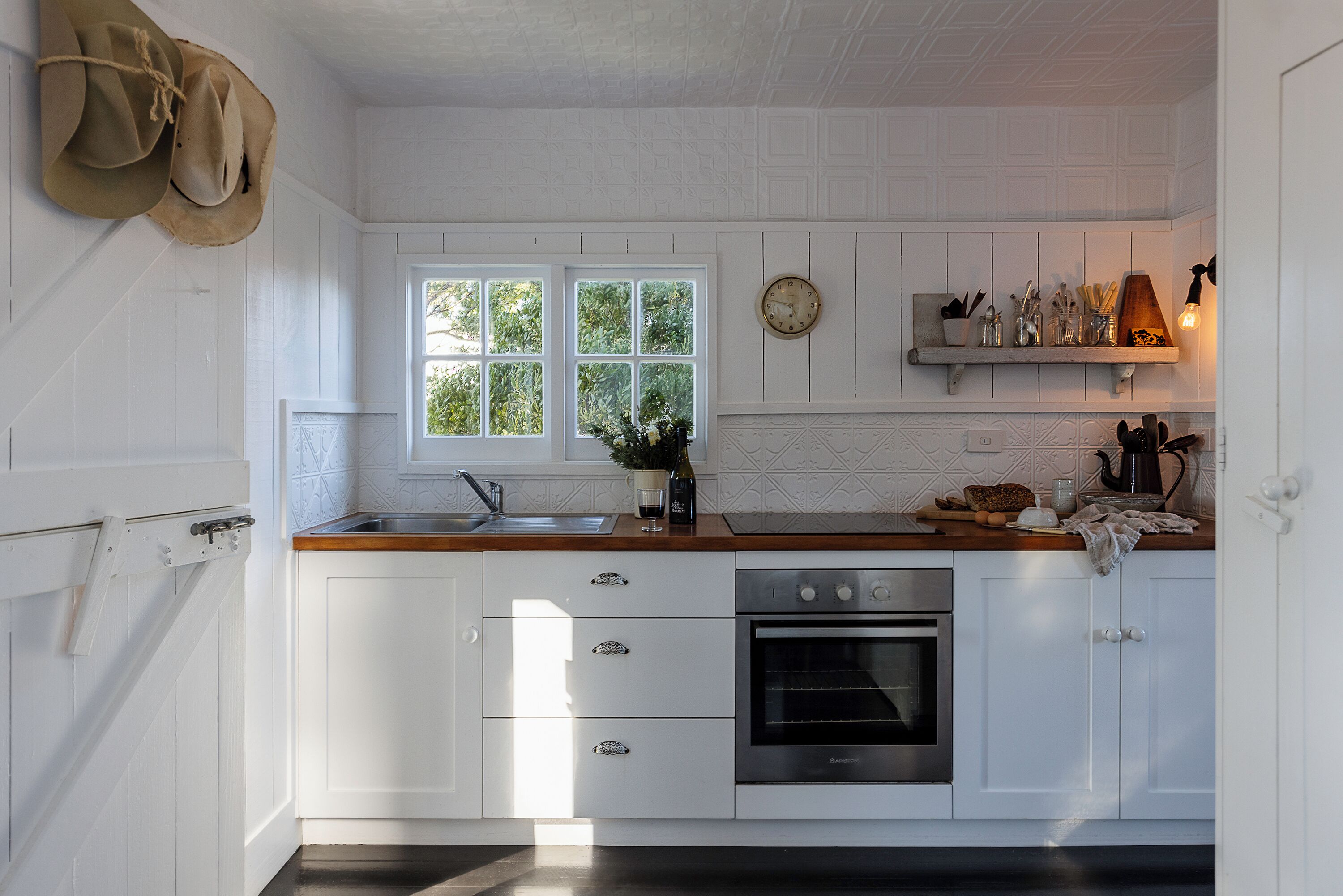 A kitchen with white cabinetry, a stainless steel oven, a window with a view of greenery, and a shelf with kitchen utensils.