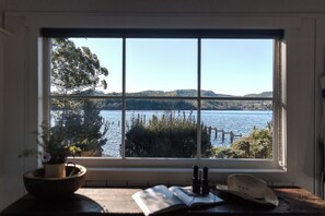 A window with a view of a lake, a book, and a hat on a table.