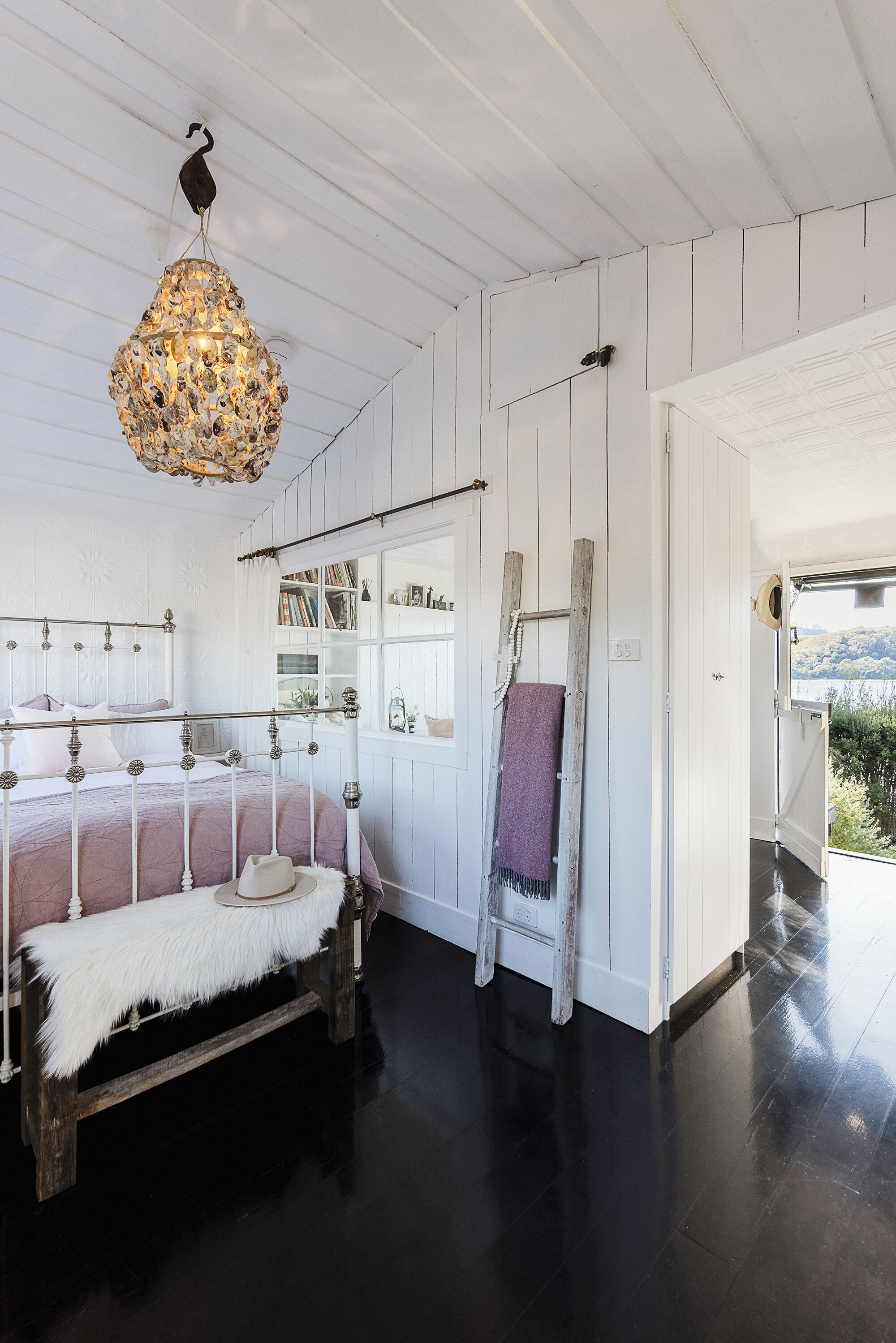 A bedroom with a bed, a wooden ladder, a hanging light fixture, and a view of the outdoors.