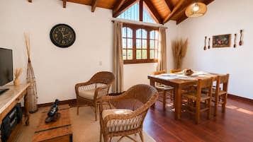 A cozy living room with a wooden ceiling, a round wall clock, wicker furniture, and a dining table set for four.