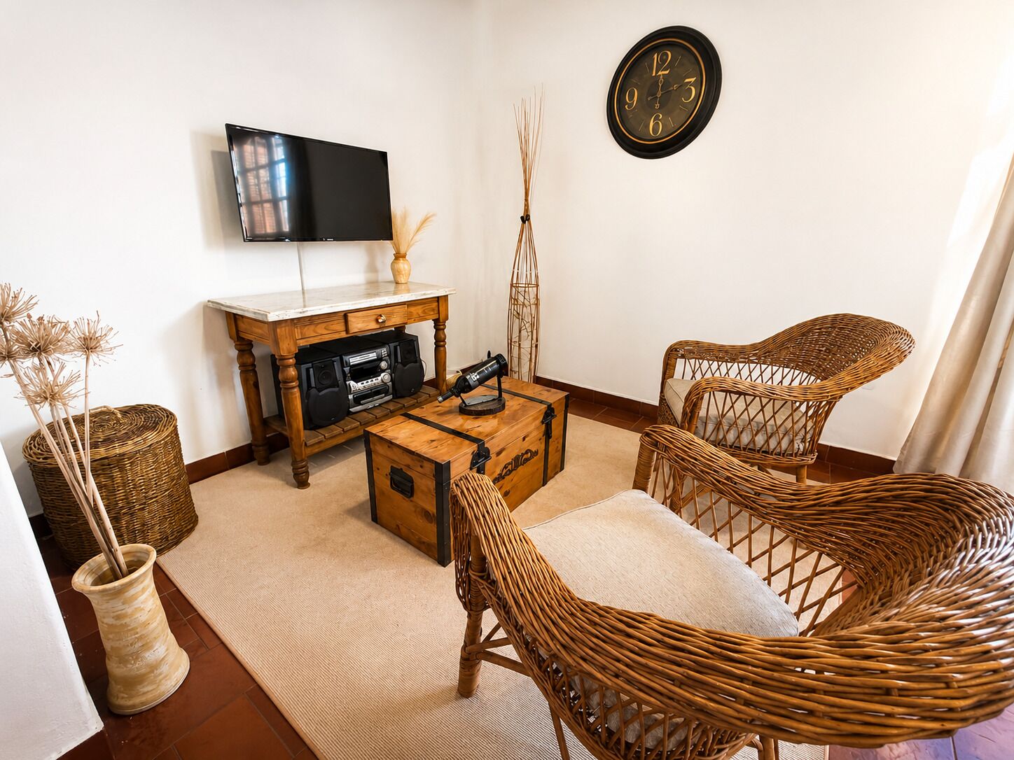 A living room with a flat-screen TV, a wooden coffee table, wicker chairs, a clock on the wall, and a decorative vase.