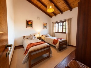 A room with two beds, a window with curtains, a hanging light fixture, and two wall-mounted paintings.