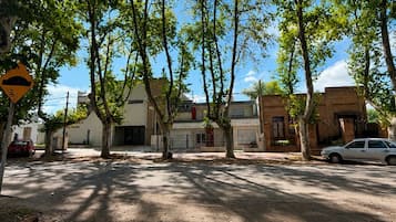A street lined with trees casting shadows on the ground, with buildings and parked cars in the background.