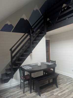 Uma sala de jantar com mesa preta, jogos americanos xadrez e um banco. Uma escadaria com corrimãos pretos é visível ao fundo.