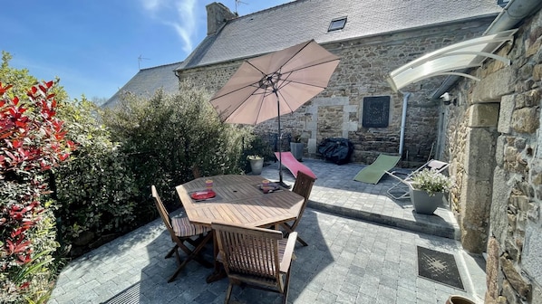Een gebouw van steen met een betegeld terras, een houten tafel met stoelen en een grote parasol.