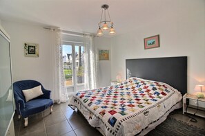 Een slaapkamer met een groot bed, een stoel, een nachtkastje met een lamp en een raam met gordijnen.