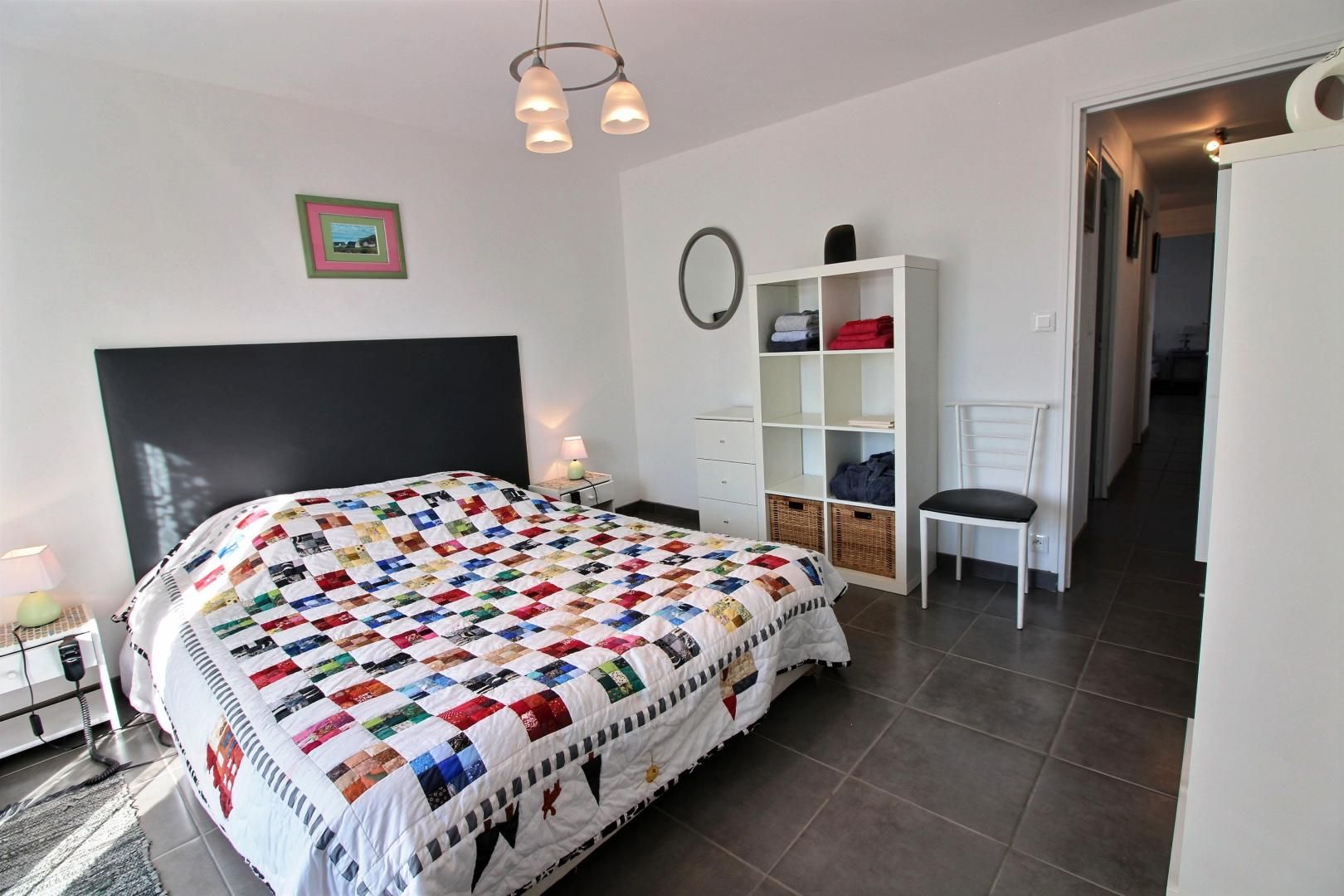 Une chambre avec un grand lit, une table de chevet avec une lampe, une armoire, une chaise et un couloir menant à une autre pièce.