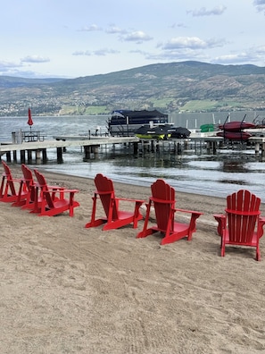 Blick auf einen See mit roten Adirondack-Stühlen, die am Sandstrand aufgereiht sind, eine Bootsanlegestelle und eine ferne Hügellandschaft.