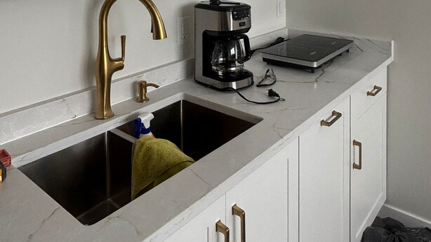 Una cocina con armarios blancos, un lavamanos de acero inoxidable, una cafetera y una ventana sobre el lavamanos.