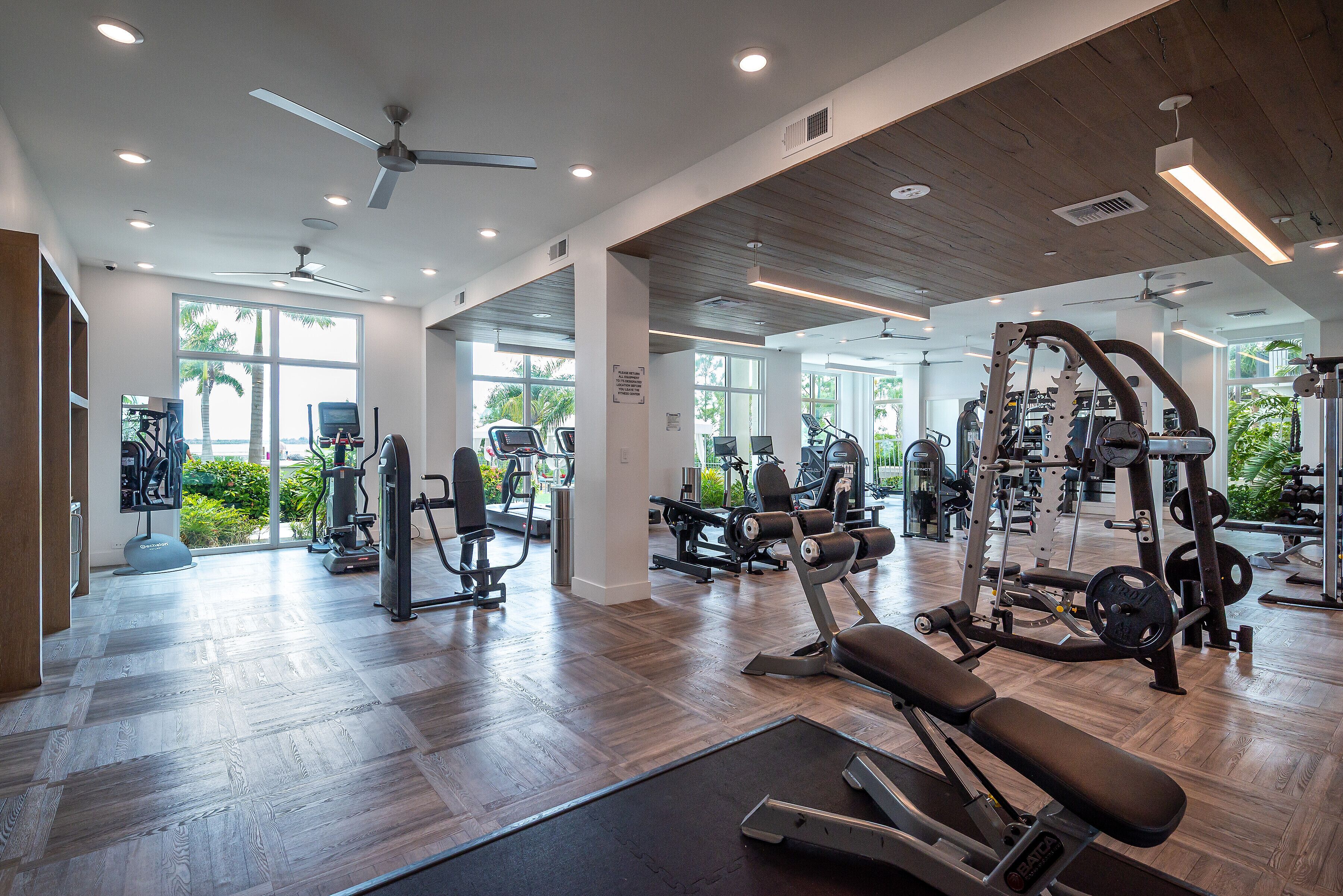 Une salle de sport moderne équipée de divers appareils d’exercice, dotée de grandes fenêtres et d’un plancher en bois.