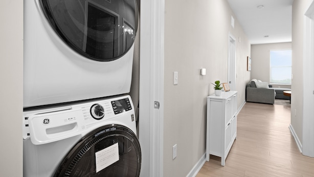 A laundry room with stacked washing machines, a vent system, and a view into a living area with a sofa and a small table. - Luxury Fragrance-Free Bayfront Retreat with Saltwater Pool (Clearwater)