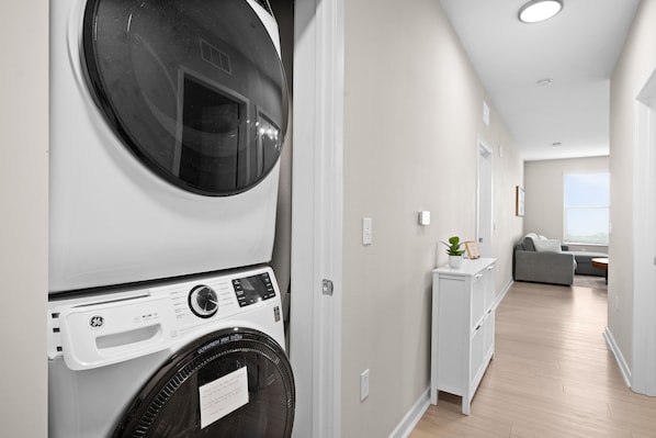 A laundry room with stacked washing machines, a vent system, and a view into a living area with a sofa and a small table.