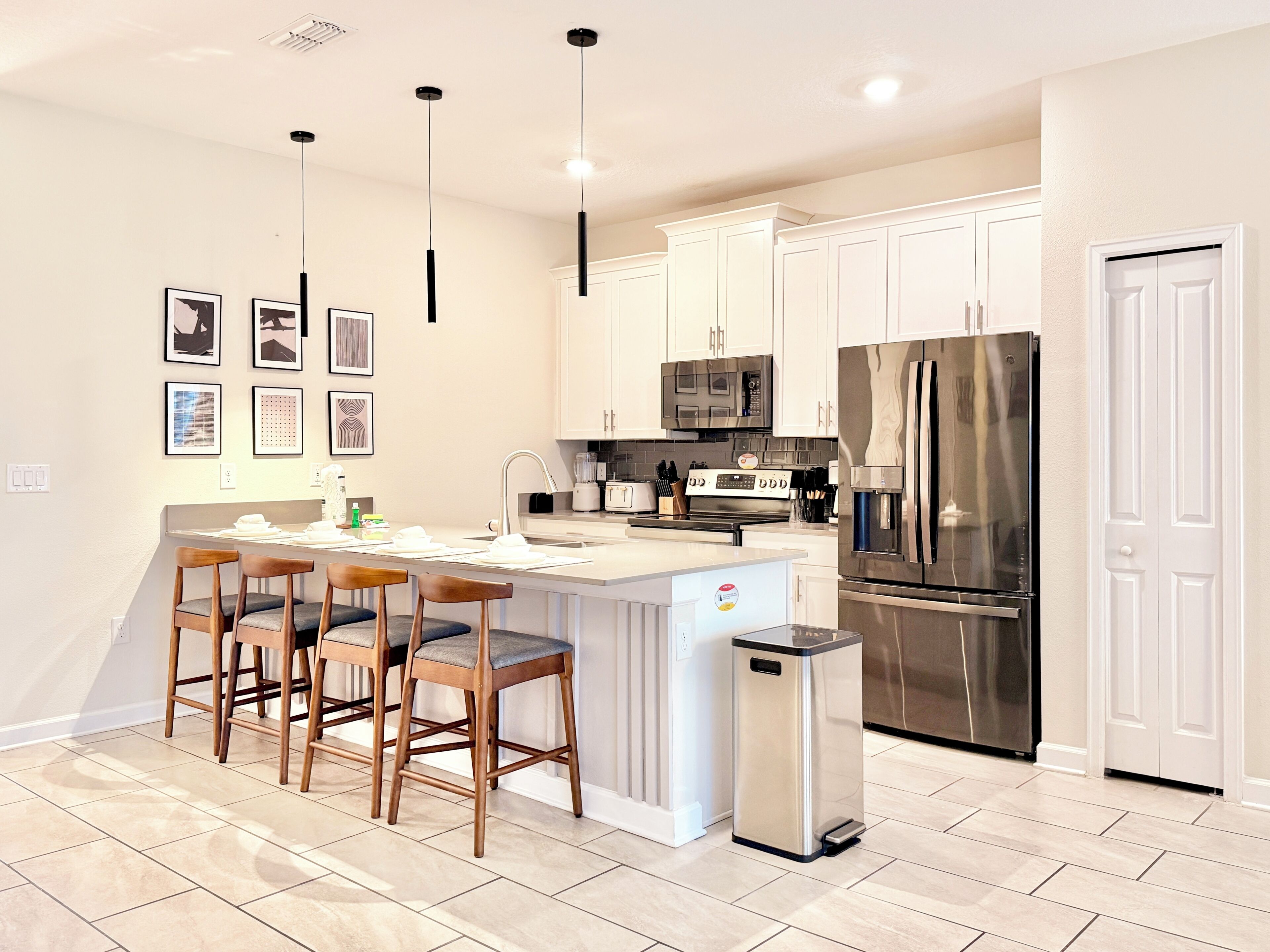 A modern kitchen with white cabinets, a stainless steel refrigerator, a built-in oven, and a bar area with stools.