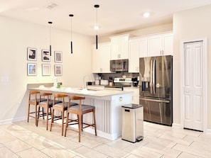 A modern kitchen with white cabinets, a stainless steel refrigerator, a built-in oven, and a bar area with stools. - Family Fun: Private Pool, Themed Rooms & Summer Kitchen - Walk to Clubhouse (Kissimmee)