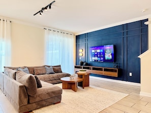 A modern living room with a sectional sofa, a coffee table, and a flat-screen TV mounted on a dark wall. - Family Fun: Private Pool, Themed Rooms & Summer Kitchen - Walk to Clubhouse (Kissimmee)