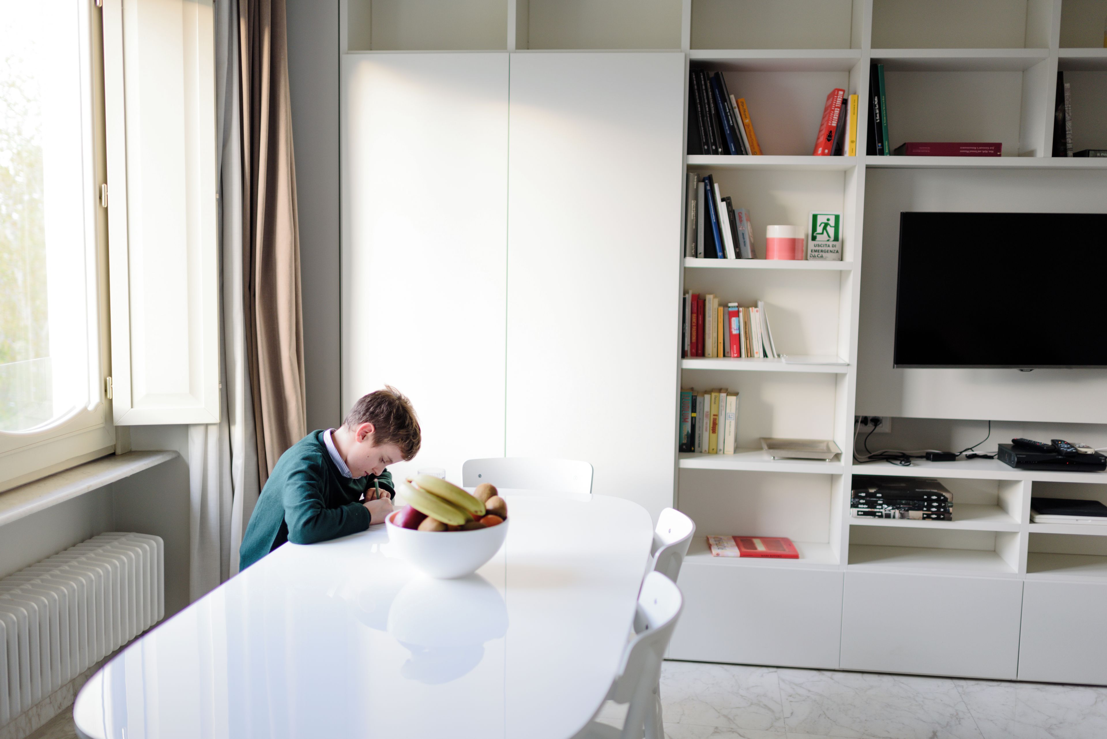 A modern living room with a white table, a person reading, a bookshelf with books, and a flat-screen TV.