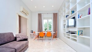 A modern living room with a sofa, dining table, chairs, and a TV on a white shelf. - Fontana Più Stella (Rome)