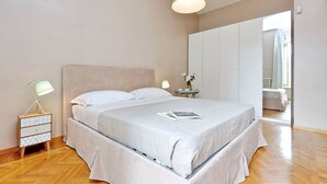 A bedroom with a bed, bedside lamps, a nightstand, a wardrobe, and a view of another room.