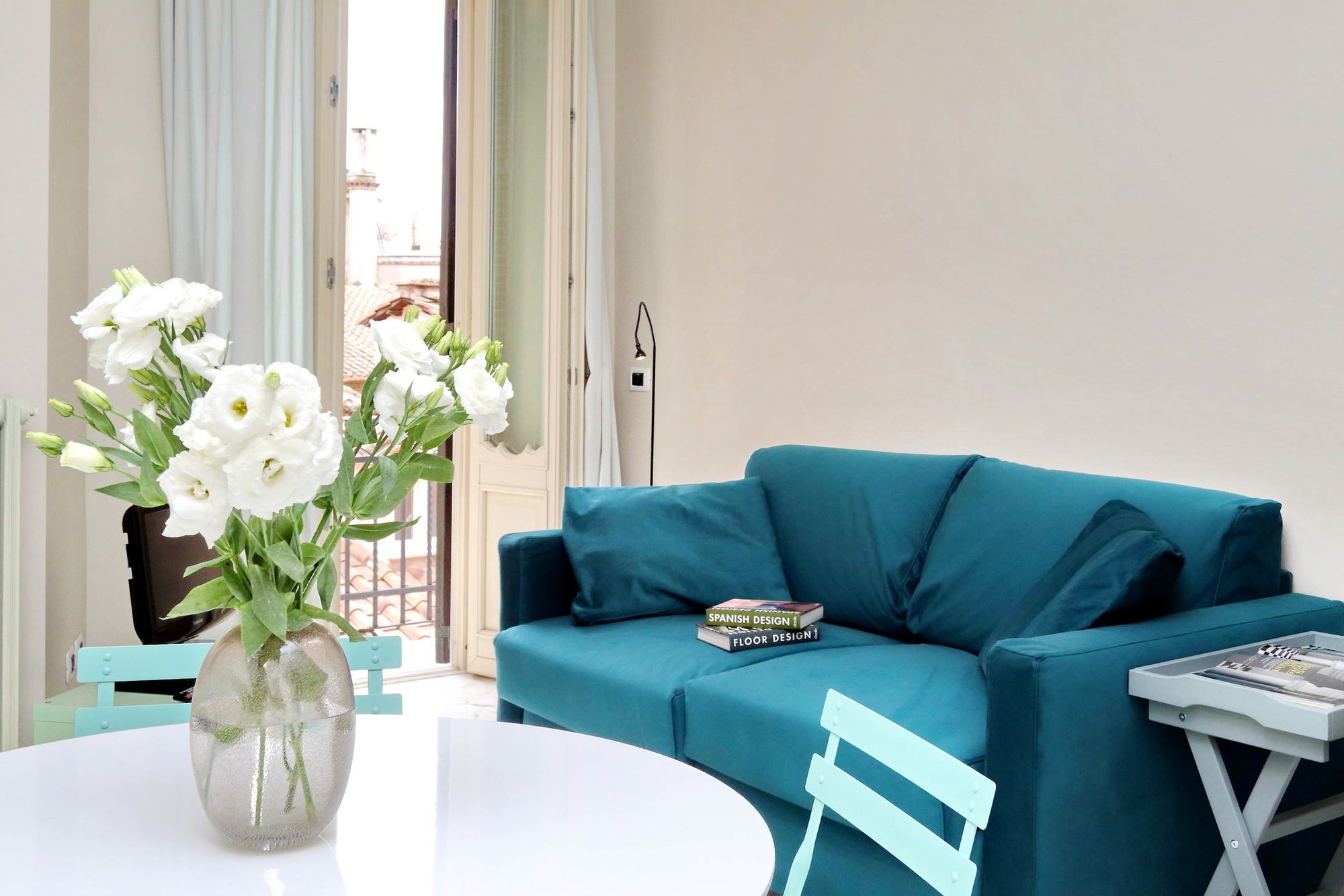 A room with a teal sofa, a white table, and a vase with white flowers.