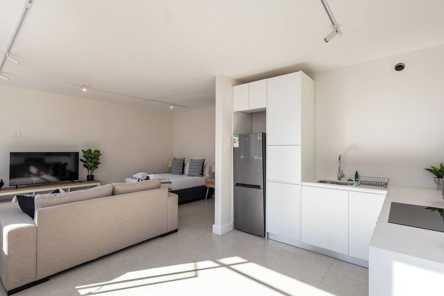 A modern living room with a sofa, a flat-screen TV, and a dining area with a refrigerator and sink.