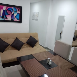 Ein Wohnzimmer mit einem beigefarbenen Sofa, schwarzen Kissen, einem Couchtisch mit einer Flasche und einem Salzstreuer, sowie einem farbenfrohen, abstrakten Gemälde an der Wand.