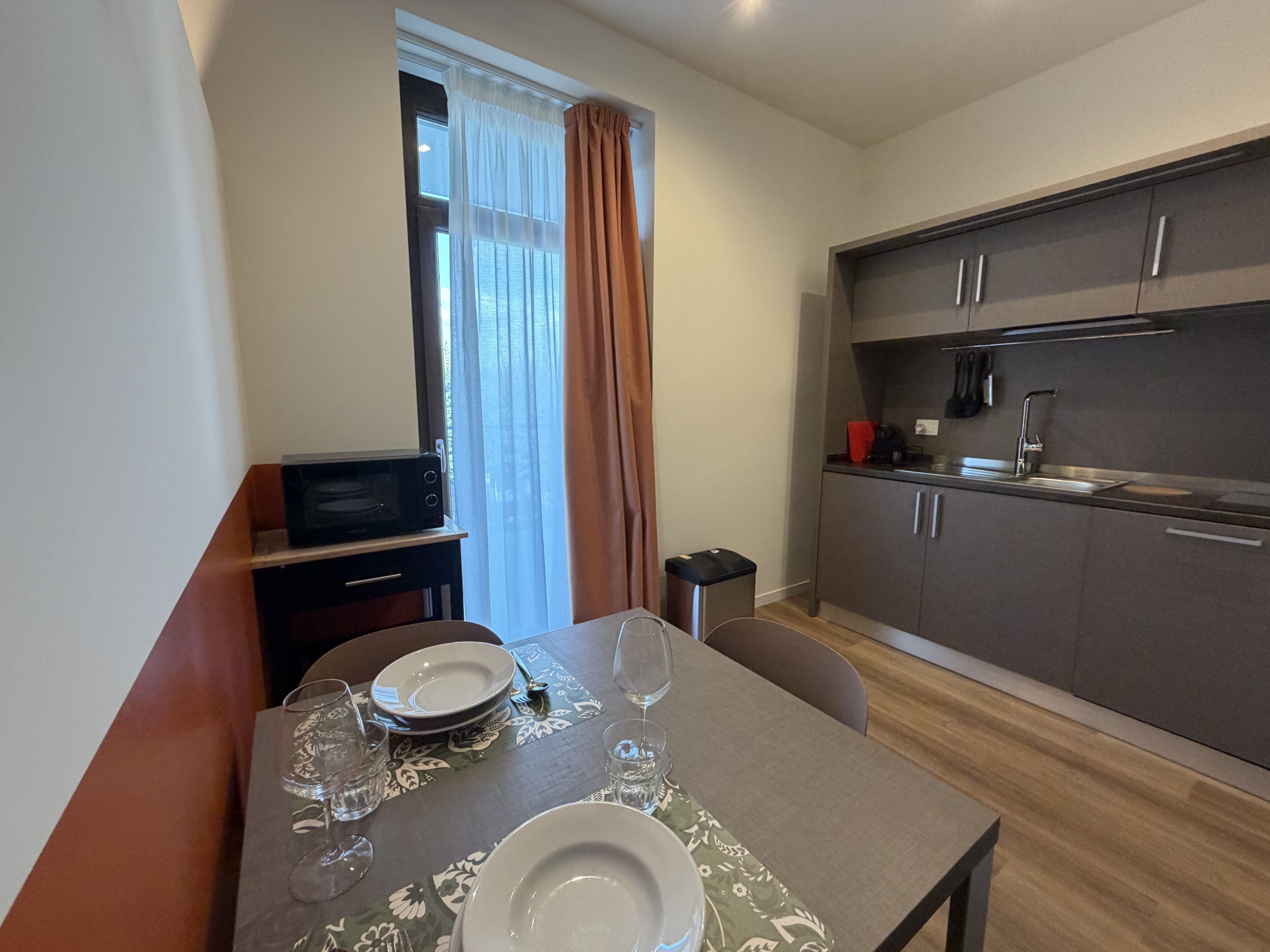 Una cucina compatta con elettrodomestici moderni, un'area pranzo con un tavolo apparecchiato per due e una finestra con tende.