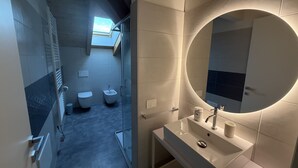 Um banheiro moderno com box de vidro, pia branca e espelho redondo, além de um claraboia sobre o vaso sanitário.