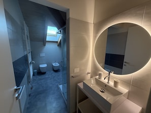 Ein modernes Badezimmer mit Duschkabine aus Glas, einem weißen Waschbecken und einem runden Spiegel sowie einem Oberlicht über der Toilette.