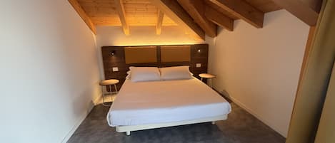 Un dormitorio con techo inclinado, una cama, mesitas de noche y un taburete pequeño.