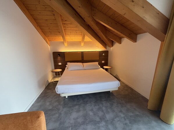Un dormitorio con techo inclinado, una cama, mesitas de noche y un taburete pequeño.
