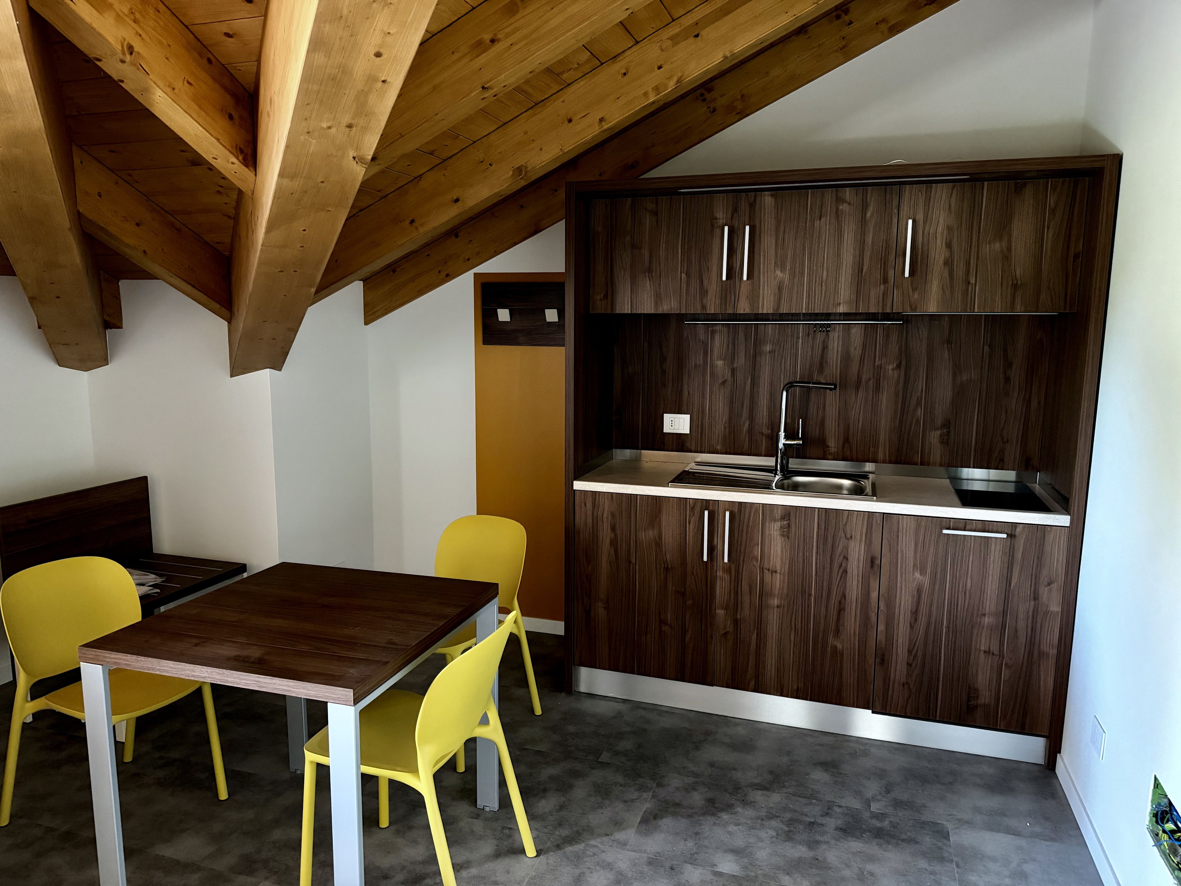 A compact kitchen with dark wooden cabinets, a stainless steel sink, and a dining area with a wooden table and yellow chairs.