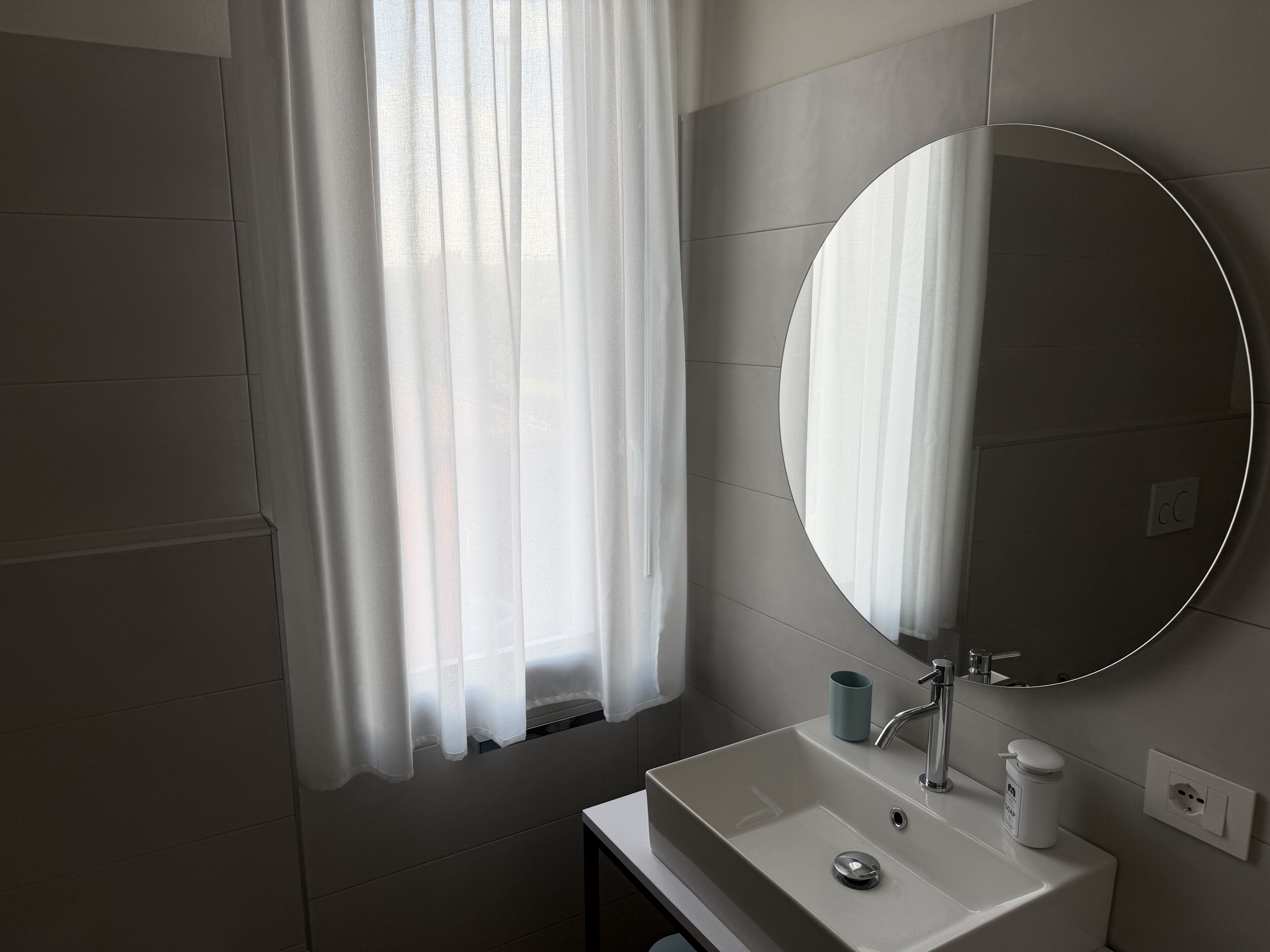 Une salle de bain avec un lavabo blanc, un miroir et une fenêtre ornée de rideaux blancs.
