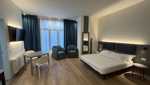 Quarto de hotel moderno com cama, escrivaninha, cadeiras e televisão.