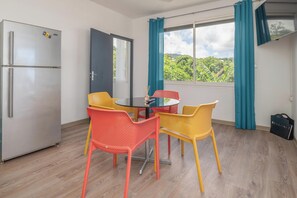 A compact kitchen with a refrigerator, a dining table with chairs, a window with curtains, and a view of greenery outside.