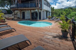 A wooden deck with a swimming pool, lounge chairs, and a building with a balcony.