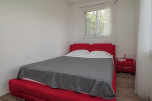 A single-bed room with a red headboard, a grey bedspread, and a small bedside table with a red lamp.