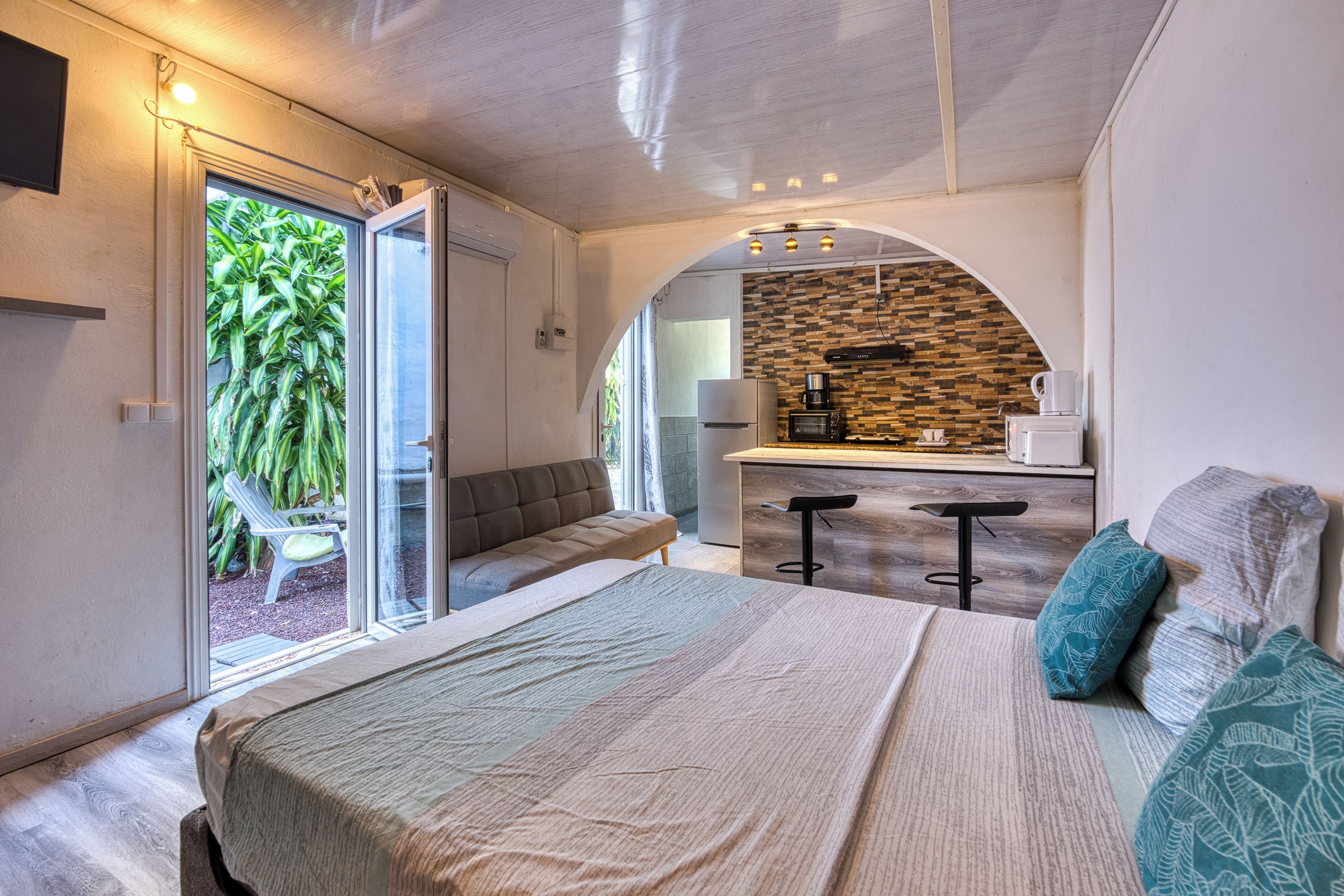 A modern bedroom with a large bed, a seating area with a bench, and a kitchenette with a stone backsplash.
