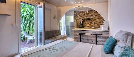 A modern bedroom with a large bed, a seating area with a bench, and a kitchenette with a stone backsplash.