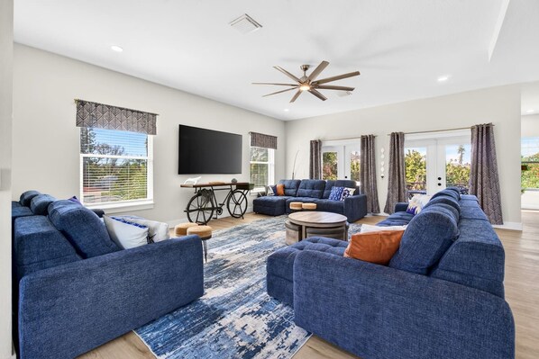 A spacious living room with a large sectional sofa, a flat-screen TV mounted on the wall, and a ceiling fan.