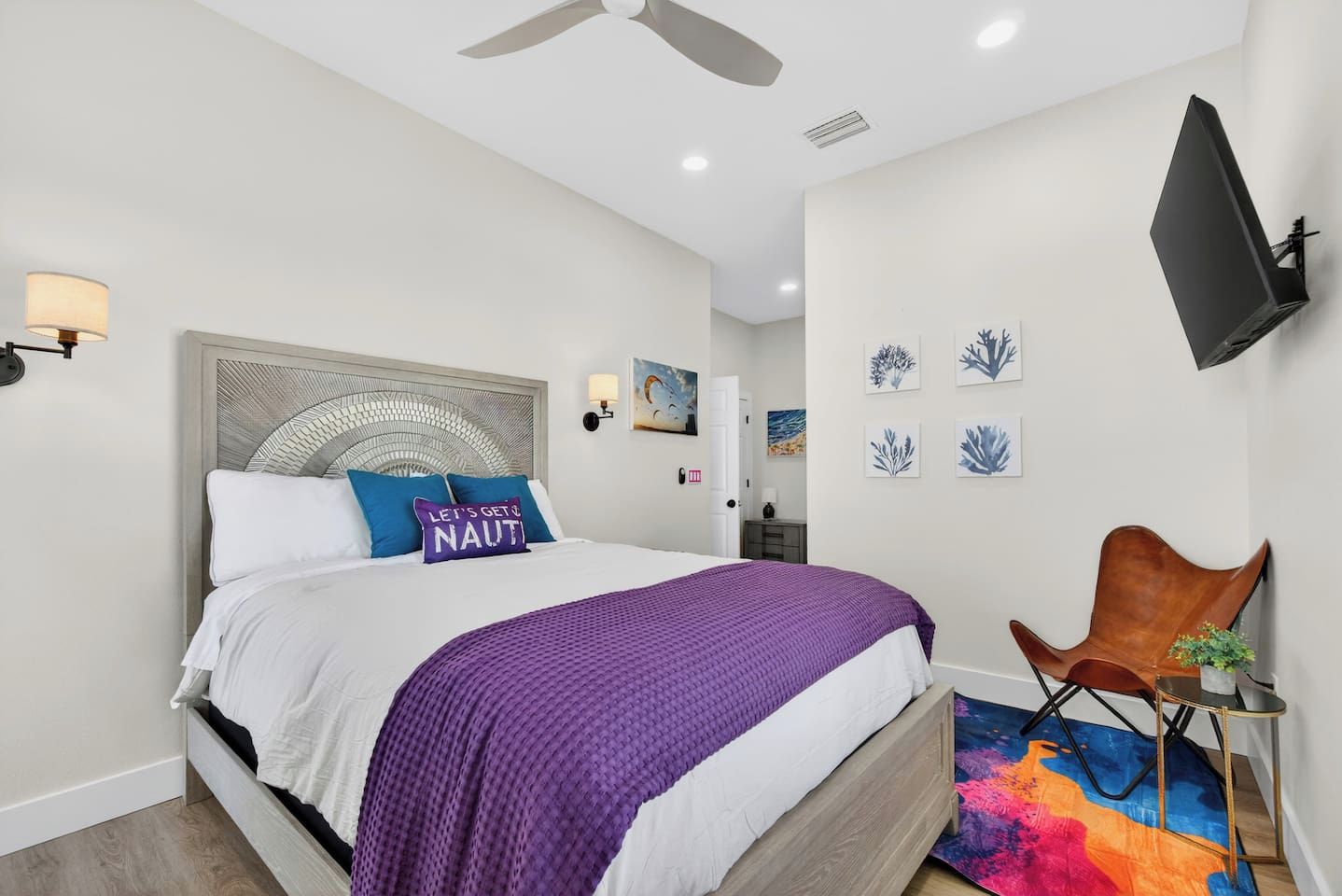 A modern bedroom with a bed, a ceiling fan, wall-mounted artwork, and a flat-screen TV.