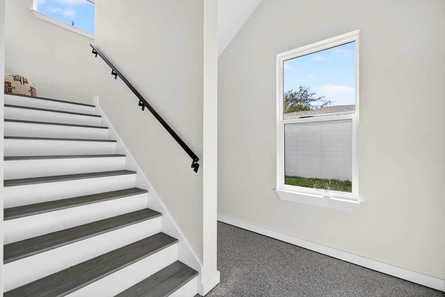 A staircase with a black handrail leading to a landing, a window with a view of a fence and trees, and a plain wall.