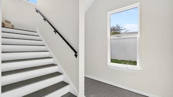 A staircase with a black handrail leading to a landing, a window with a view of a fence and trees, and a plain wall.
