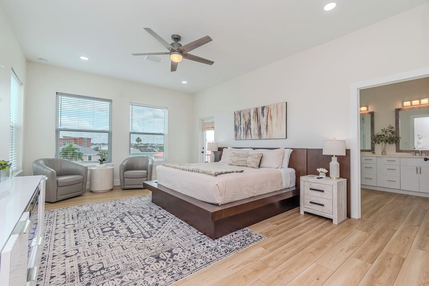 A modern bedroom with a large bed, two bedside tables, a ceiling fan, and a view of the city through large windows.