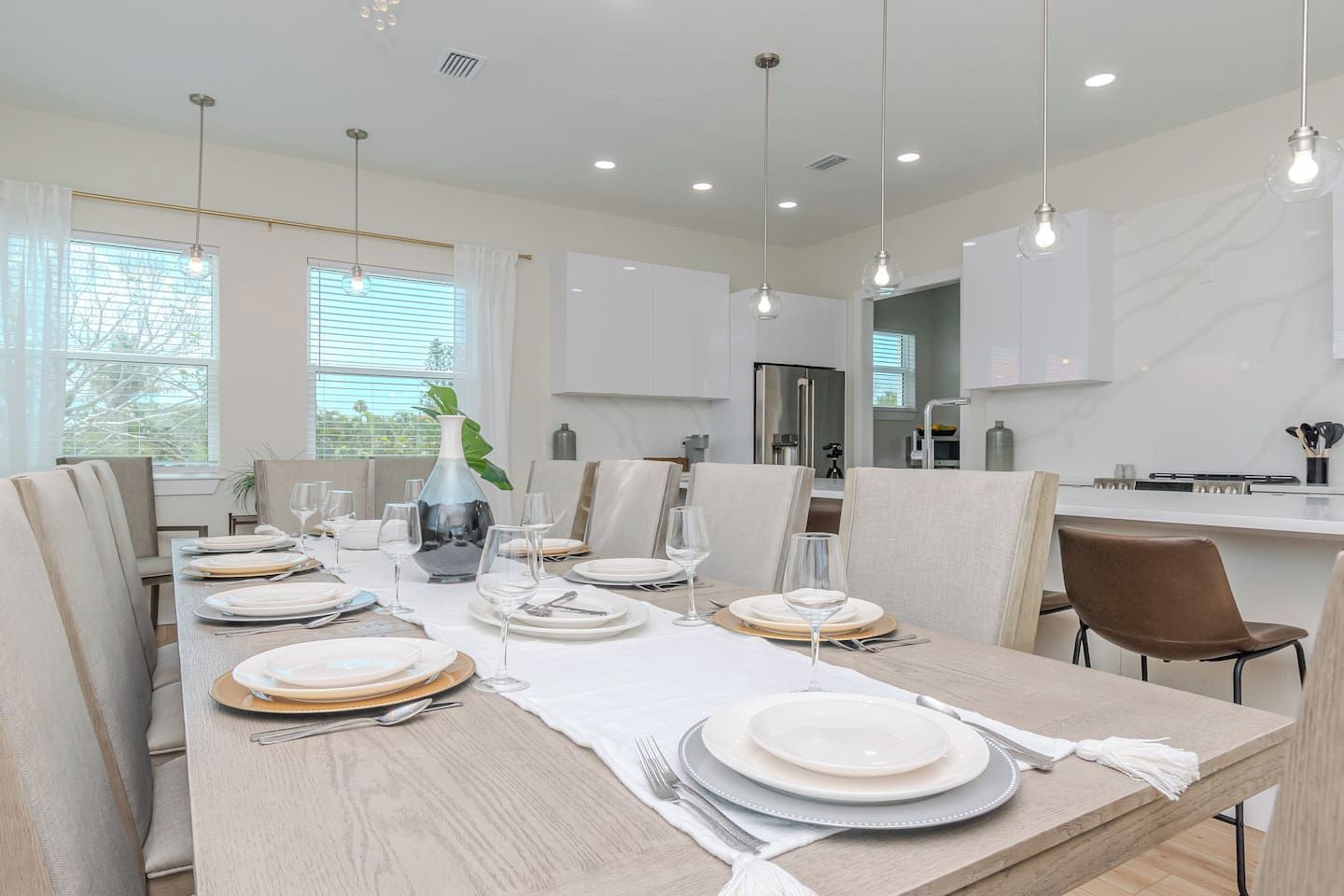A modern dining area with a long table set for a meal, surrounded by chairs, and a kitchen in the background.