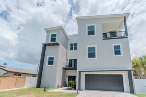 A modern multi-story residential building with a garage, a balcony on the upper floor, and a fenced yard.